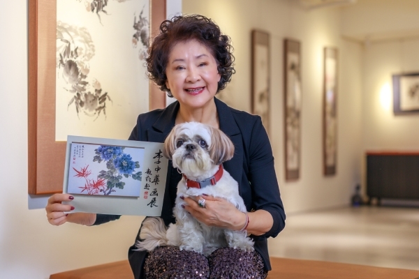 李中華一手抱著愛犬，一手拿著昔日開畫展時印製的宣傳卡，在胡克敏紀念館拍照。（圖片來源：葉琳喬攝）