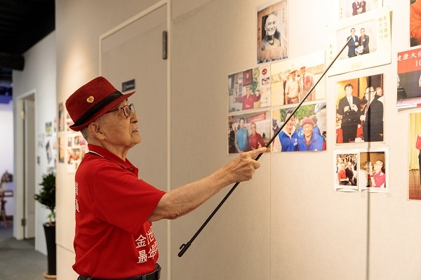 林友茂右手拿指揮棒，腰桿挺直，解說騷貓公益咖啡的照片牆。（圖片來源：葉琳喬攝 場地協力：騷貓公益咖啡）